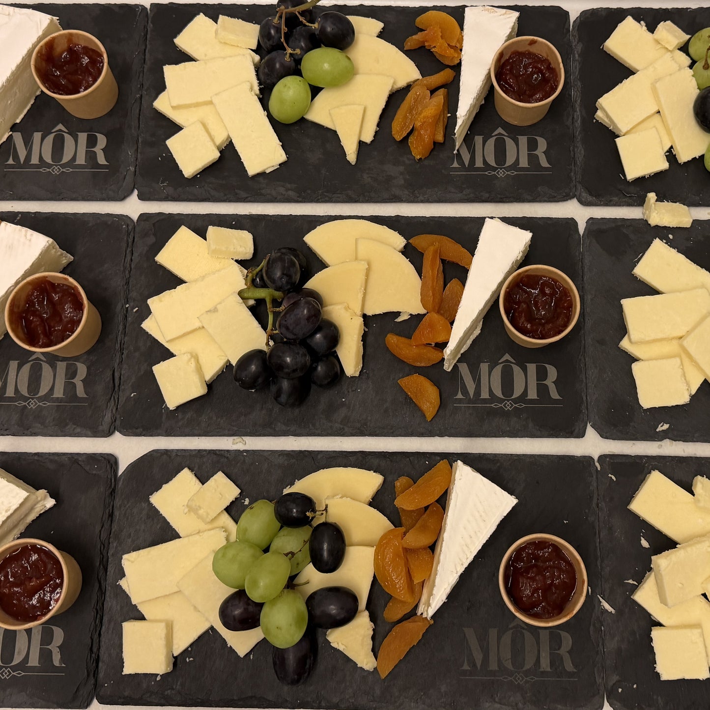 Multiple cheese platters with cheese, grapes, dried fruits, and jam on black slate boards with 'Mör' branding for a wine tasting.