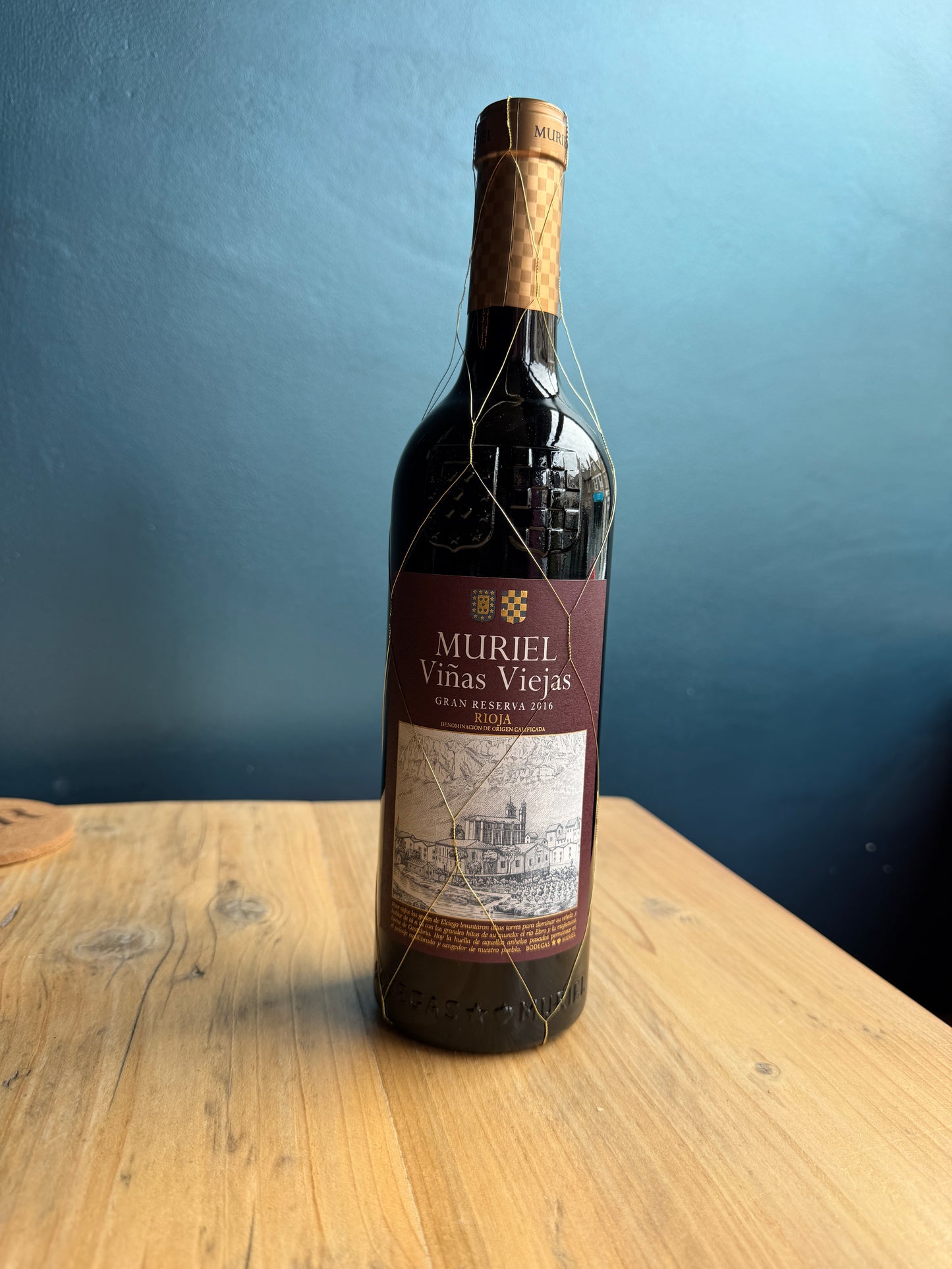 A bottle of Muriel Gran Reserva Rioja red wine on a light-colored wooden table against a dark blue background.