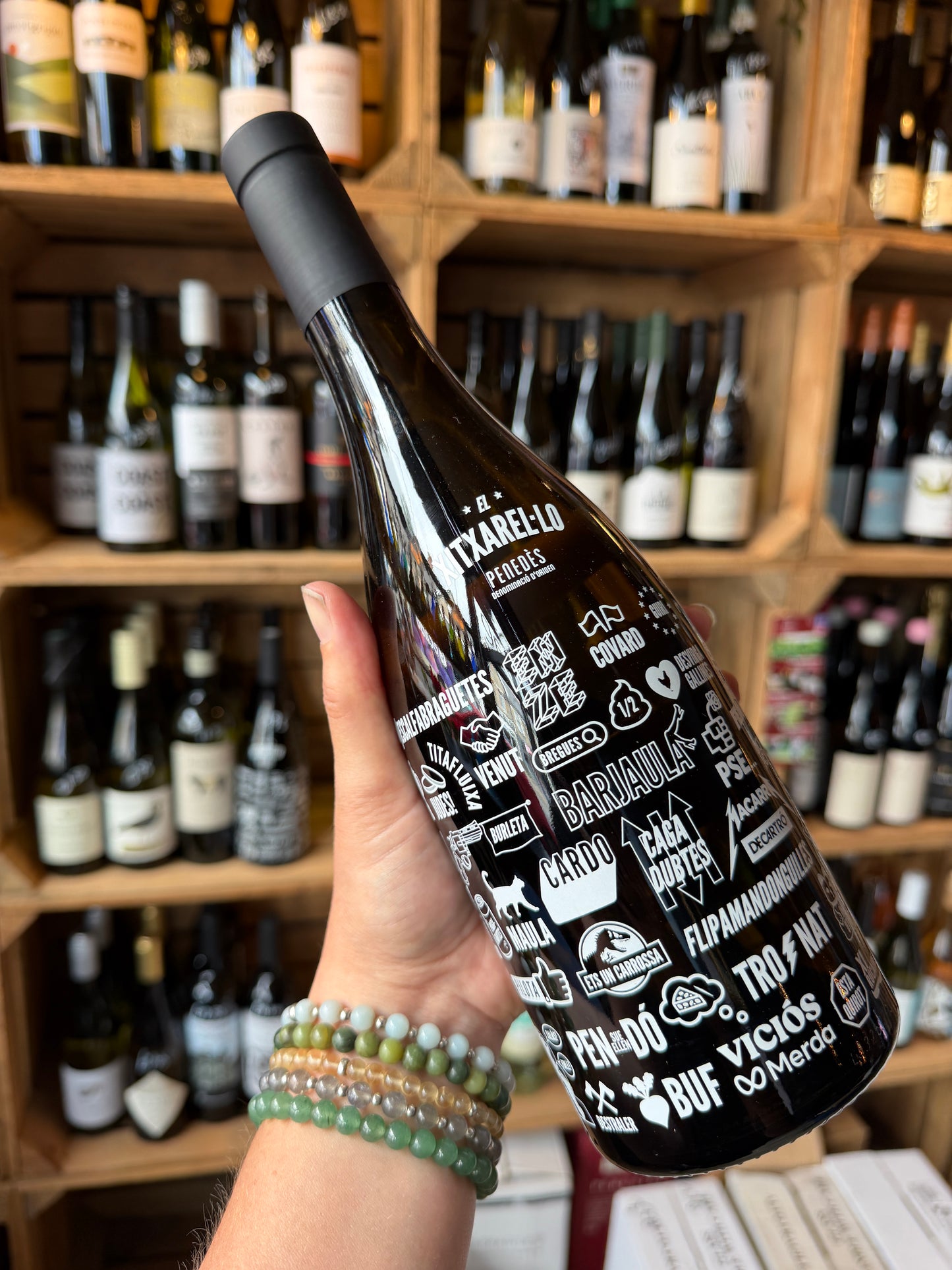 A hand holding a bottle of Can Virgili El Xitxarel·lo, set against a background of wooden wine shelves filled with other bottles in Mor wine bar and wine shop in Barry South Wales.
