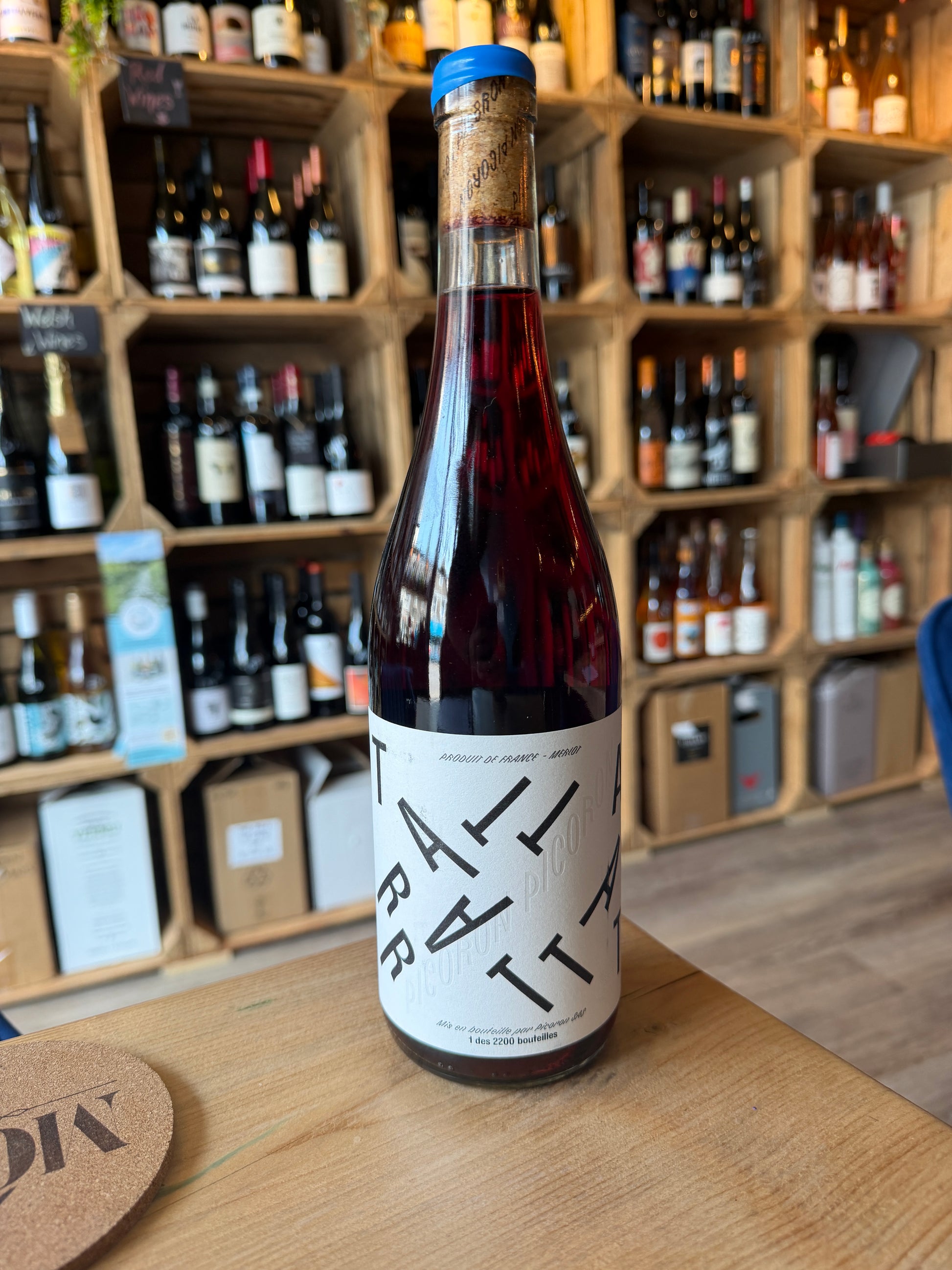 A bottle of Château Picoron Tattarrattat Carbonic Maceration Merlot Chillable Natural Red on a wooden table in front of a wine shop display of shelves filled with bottles in a wine shop in Barry