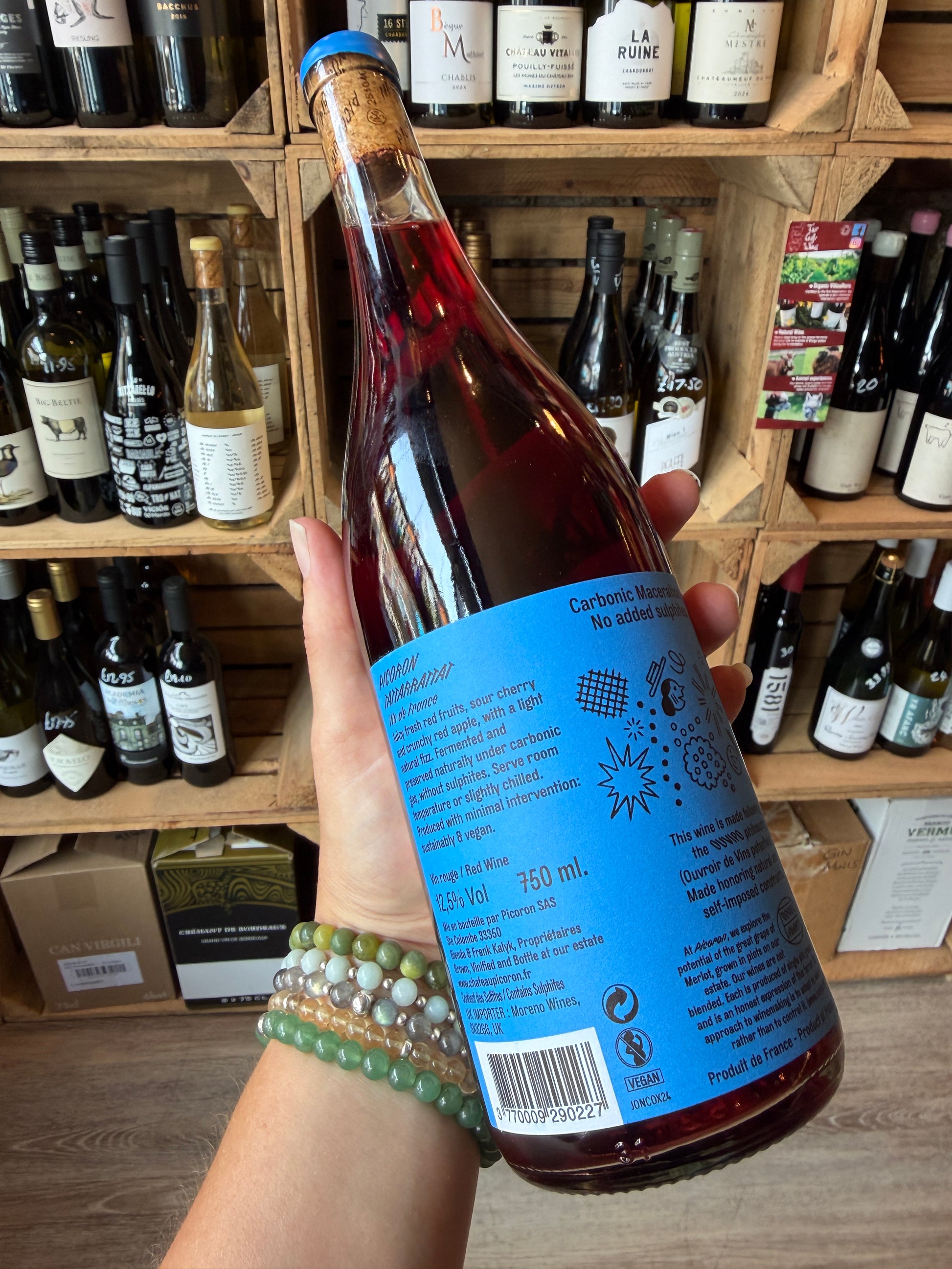 A hand holding a bottle of Château Picoron Tattarrattat Carbonic Maceration Merlot (Chillable Natural Red) showing the back label set against a background of wooden wine racks filled with other wine bottles.