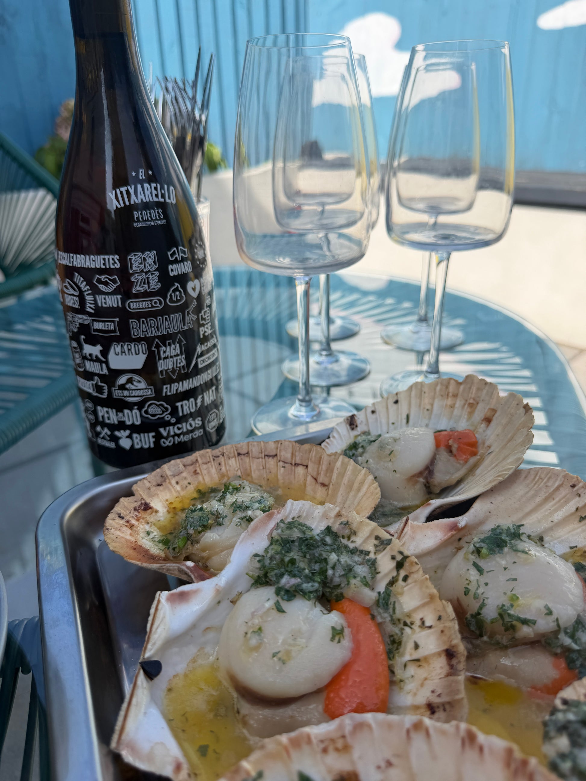 A bottle of El Xitxarel-lo with a tray of BBQ cooked scallops topped with herbs and garlic, alongside wine glasses on a table outdoors on a sunny patio.