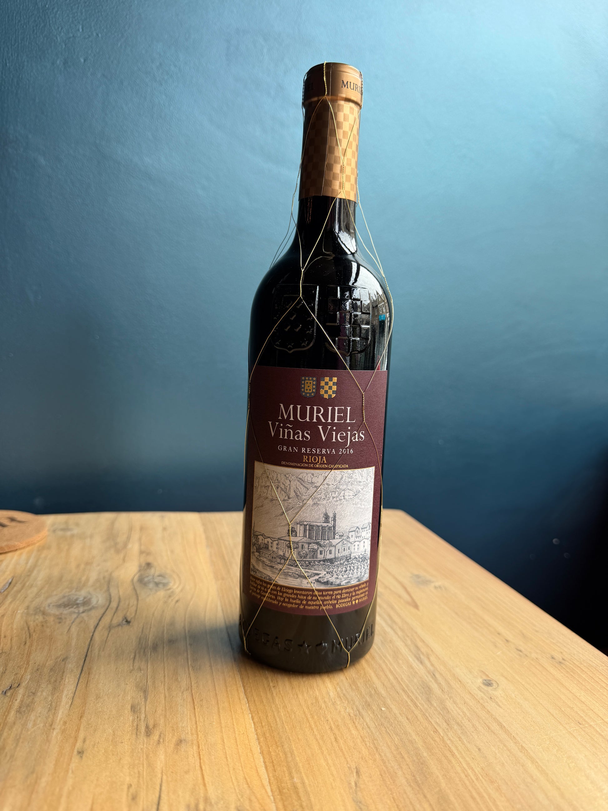 A bottle of Muriel Gran Reserva Rioja red wine on a light-colored wooden table against a dark blue background.